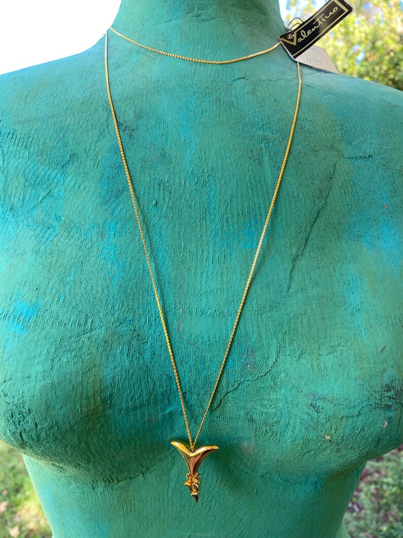 May include: A gold chain necklace with a gold shark fin pendant. The necklace is on a mannequin with a teal blue torso. The tag on the necklace reads "alexine".