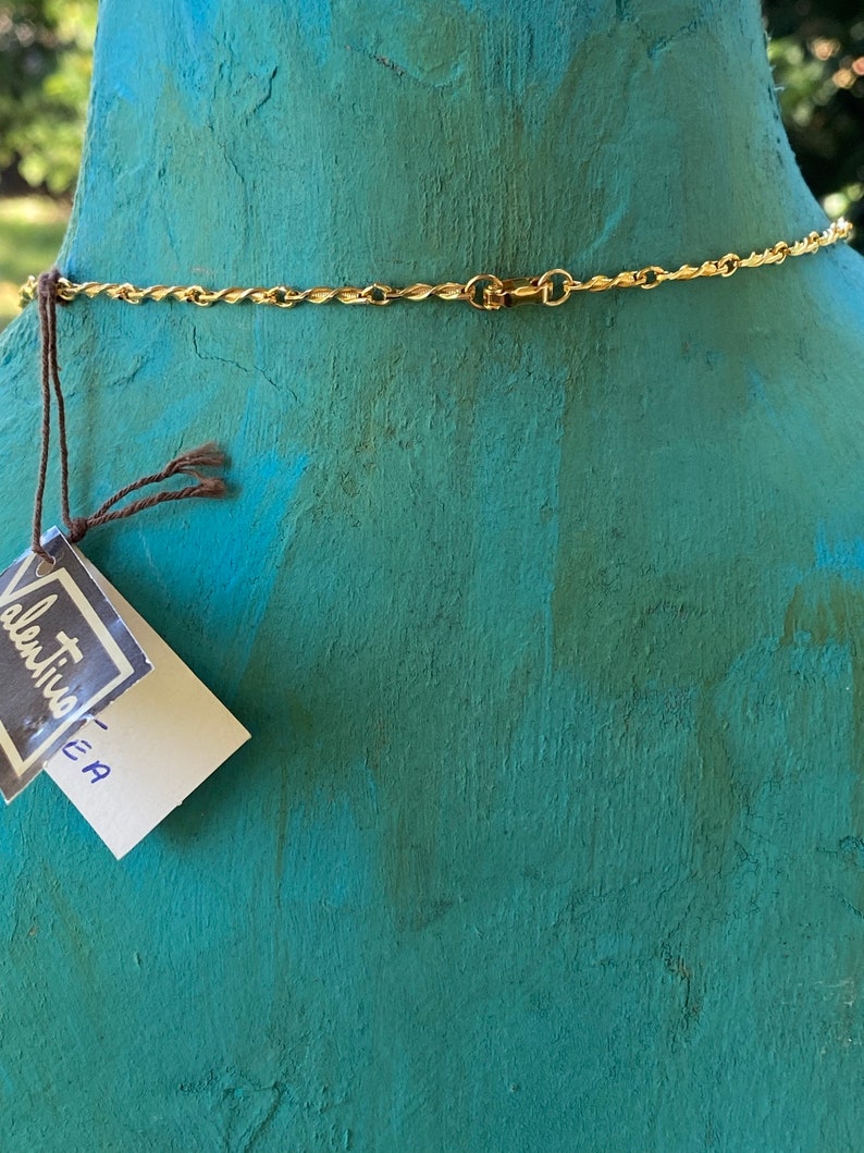 Puede incluir: Vista posterior de un collar de cadena dorada con cierre. El collar est&aacute; en un maniqu&iacute; turquesa. Una peque&ntilde;a etiqueta blanca con el texto "Valentino - EA" est&aacute; unida al maniqu&iacute;.