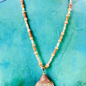 May include: A necklace with a seashell pendant. The necklace is made of brown, white, and gold beads. The seashell is a light brown color with a gold trim.