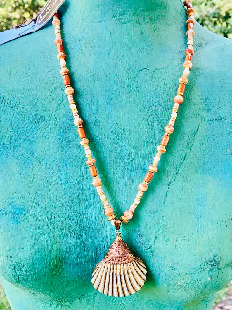 May include: A necklace with a seashell pendant. The necklace is made of brown, white, and gold beads. The seashell is a light brown color with a gold trim.