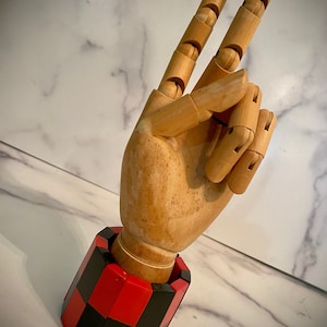 May include: A wooden articulated hand displaying the peace sign, with two fingers extended. The hand is attached to a red and black checkered base. The hand is made of light-colored wood.