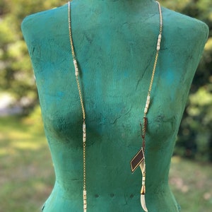 Puede incluir: Un collar largo de cadena dorada con cuentas de cer&aacute;mica blanca y detalles dorados. El collar tiene dos colgantes blancos largos al final.