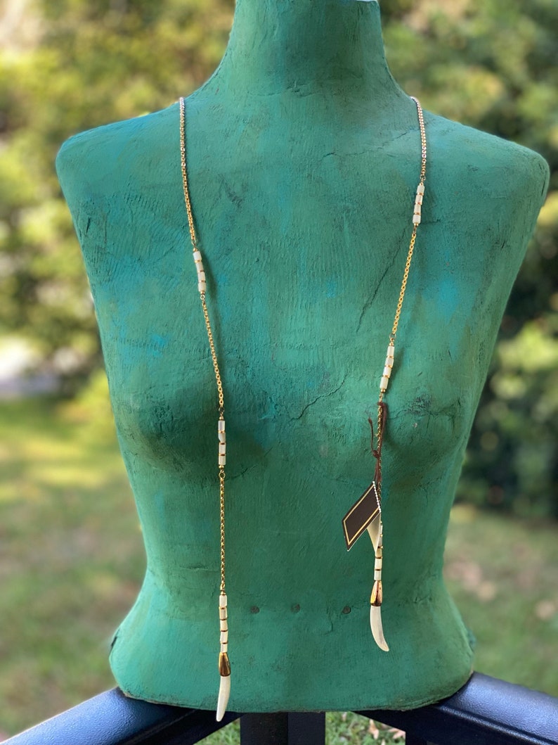 Puede incluir: Un collar largo de cadena dorada con cuentas de cer&aacute;mica blanca y detalles dorados. El collar tiene dos colgantes blancos largos al final.
