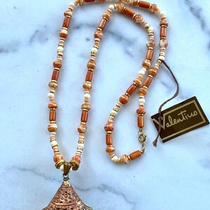 May include: A long necklace with a shell pendant. The necklace is made of brown and white beads with gold accents. The shell pendant is white with a copper-colored design. The necklace comes with a Valentino tag.
