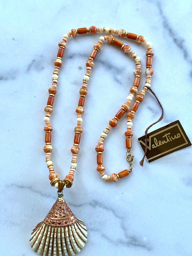 May include: A long necklace with a shell pendant. The necklace is made of brown and white beads with gold accents. The shell pendant is white with a copper-colored design. The necklace comes with a Valentino tag.