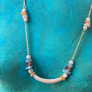 Puede incluir: Un collar de cadena dorada con un colgante de concha blanca y cuentas de concha multicolores. Las cuentas son en tonos de marr&oacute;n, verde y blanco.