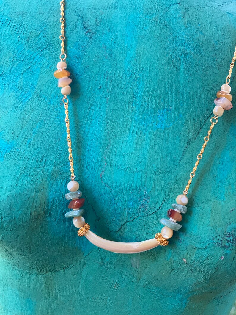 Puede incluir: Un collar de cadena dorada con un colgante de concha blanca y cuentas de concha multicolores. Las cuentas son en tonos de marr&oacute;n, verde y blanco.