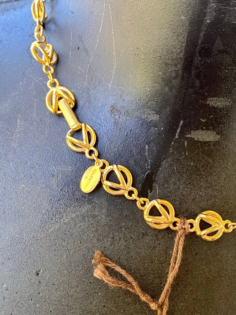 May include: A gold-toned necklace with a chain made of interlocking circles. The necklace has a small oval tag with the word "VALENTINO" inscribed on it.