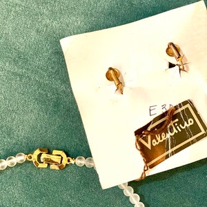 Puede incluir: Un collar de cuentas de vidrio blanco con un cierre dorado y una etiqueta de Valentino. El collar est&aacute; sobre una superficie verde.