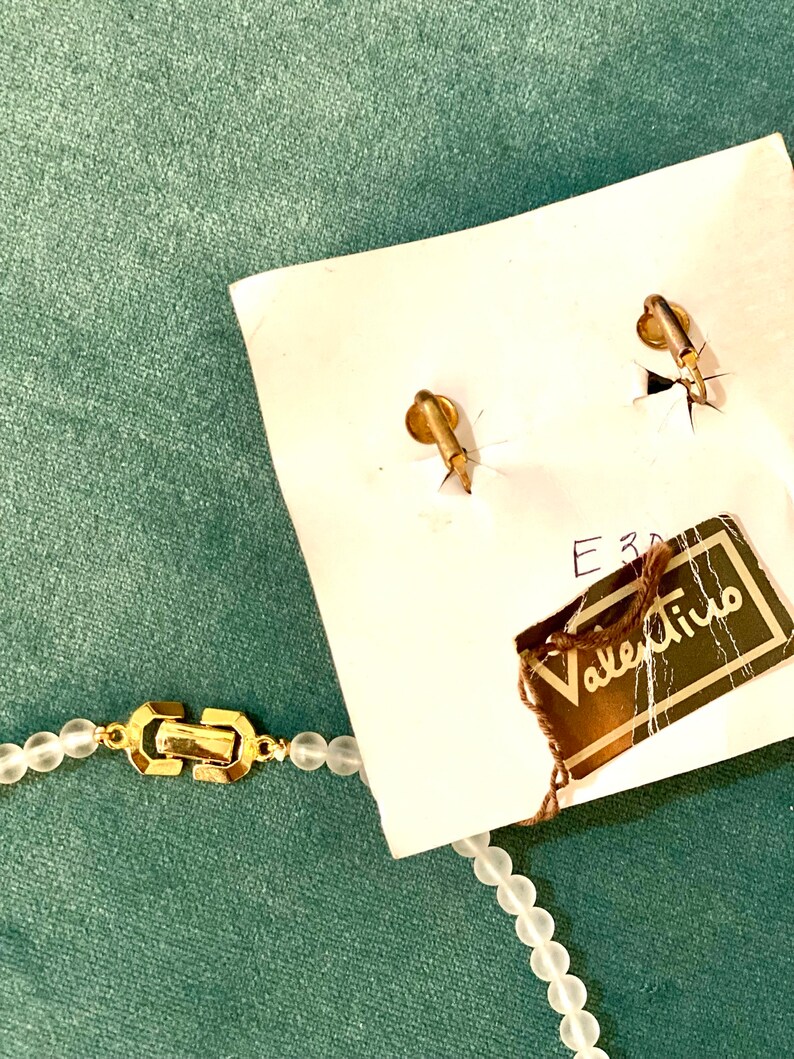 Puede incluir: Un collar de cuentas de vidrio blanco con un cierre dorado y una etiqueta de Valentino. El collar est&aacute; sobre una superficie verde.