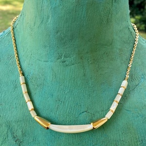 Puede incluir: Un collar de cadena dorada con cuentas de concha blanca y un acento dorado. El collar est&aacute; en un maniqu&iacute;.