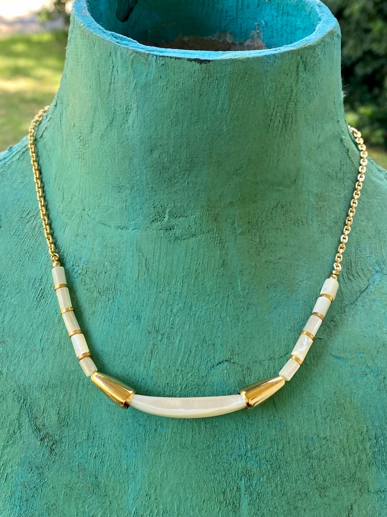 Puede incluir: Un collar de cadena dorada con cuentas de concha blanca y un acento dorado. El collar est&aacute; en un maniqu&iacute;.