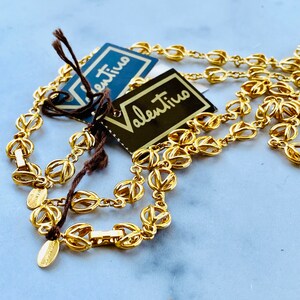 May include: A gold-toned chain necklace with a delicate, intricate design. The necklace is layered and features two tags with the word "Valentino" written in black.
