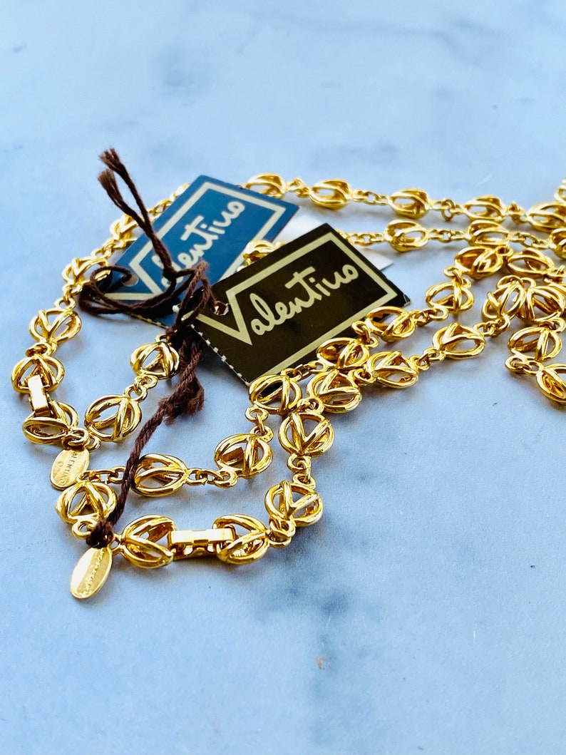 May include: A gold-toned chain necklace with a delicate, intricate design. The necklace is layered and features two tags with the word "Valentino" written in black.