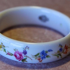 May include: White porcelain bangle bracelet with a floral design. The flowers are in shades of pink, blue, yellow, and green.