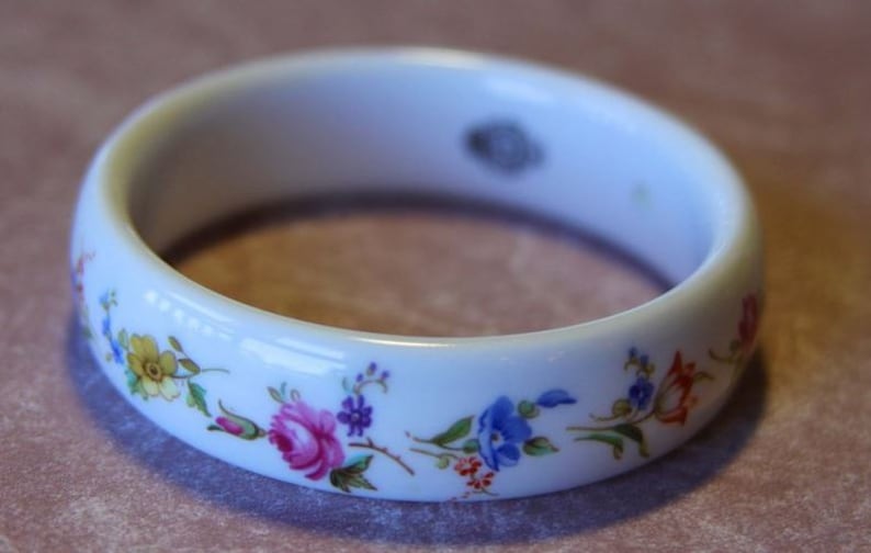 May include: White porcelain bangle bracelet with a floral design. The flowers are in shades of pink, blue, yellow, and green.
