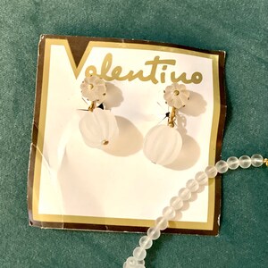 Puede incluir: Un par de pendientes de cristal blanco en forma de flor con detalles dorados en una tarjeta blanca con la palabra "valentino" impresa en ella. Tambi&eacute;n se puede ver un collar de cuentas blancas con detalles dorados en la imagen.