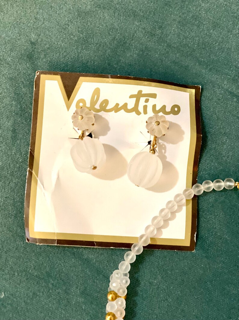 Puede incluir: Un par de pendientes de cristal blanco en forma de flor con detalles dorados en una tarjeta blanca con la palabra "valentino" impresa en ella. Tambi&eacute;n se puede ver un collar de cuentas blancas con detalles dorados en la imagen.