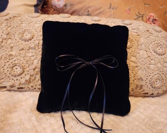 May include: A black velvet ring pillow with a satin ribbon tied in a bow. The pillow is sitting on a white crocheted blanket.