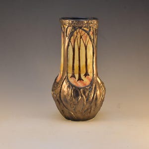 May include: A decorative vase with a bronze-colored exterior and a painted interior scene of trees. The vase has a bulbous base and a slender neck, with a detailed, textured design. The interior features yellow, orange, and brown colors.