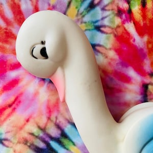 May include: A white swan figurine with a smooth matte finish, featuring a curved neck, a soft pink beak, and black painted eyes with long eyelashes. The swan has a light blue gradient on its lower body and wings, captured in a side profile view.