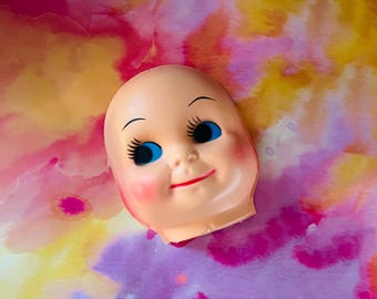 May include: A plastic doll head featuring a smooth, pale peach-colored surface, bright blue eyes with long black painted eyelashes, arched eyebrows, and a small red mouth. The face has soft pink blush on the cheeks and a rounded, bald forehead.