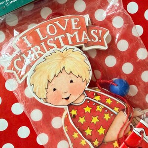 May include: A flat, die-cut Christmas ornament featuring a cartoon child with blonde hair wearing red pajamas with yellow stars. The ornament has a banner at the top with the text I LOVE CHRISTMAS! and includes a small blue bead and red string.