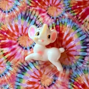 May include: A small, white plastic elephant figurine with a glossy finish. The elephant has large ears with pink inner accents, black painted eyes with long eyelashes, a small pink mouth, and a light pink patch on its chest, depicted in a playful pose.