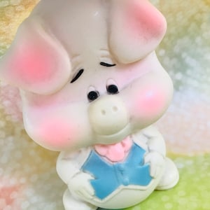 May include: A white pig figurine with pink cheeks and a blue and pink bow tie. The pig has a sweet expression and is looking to the right.