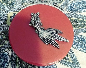 May include: A red circular pendant with a silver fish design. The pendant is attached to a silver chain.