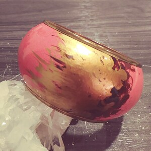 May include: A pink and gold bangle bracelet with a metallic finish. The bracelet has a wide band with abstract gold and black paint splatters.