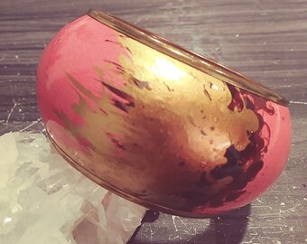 May include: A pink and gold bangle bracelet with a metallic finish. The bracelet has a wide band with abstract gold and black paint splatters.