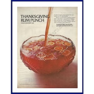 May include: An advertisement for Thanksgiving Rum Punch. A clear glass punch bowl is filled with a red liquid and orange slices. A stream of liquid is being poured into the bowl. The text reads "Thanksgiving Rum Punch".