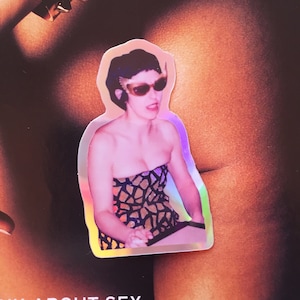 Pode incluir: Um adesivo com uma mulher vestindo um vestido estampado em preto e marrom e óculos de sol. O adesivo tem um acabamento holográfico.