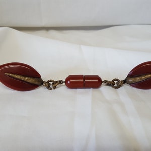 May include: A pair of red, leaf-shaped, plastic, decorative clasps connected by a gold-toned chain. The clasps have a gold-toned design on the front.