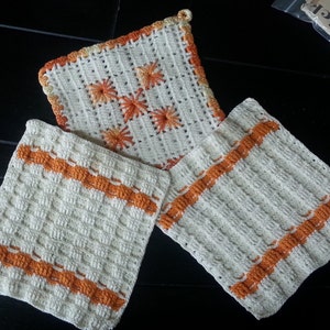 Könnte beinhalten: Drei gehäkelte Topflappen in Weiß mit orangefarbenen Akzenten. Die Topflappen sind mit einem einfachen Stichmuster gearbeitet und haben eine quadratische Form.
