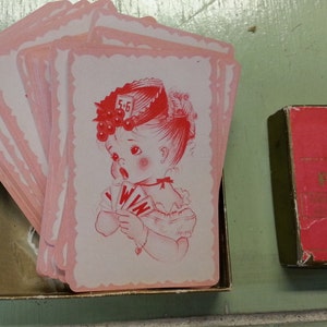 Puede incluir: Una baraja de cartas de juego de color rosa con una ilustración roja y blanca de una niña que lleva un sombrero rojo y sostiene una carta que dice "WIN". La caja de las cartas es roja y dice "Fun With Words! Playing Cards".