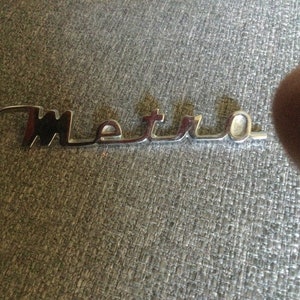 以下が含まれることがあります： 「Metro」という文字が浮き彫りになった銀色の金属製のロゴです。