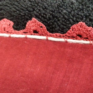 Puede incluir: Primer plano de una tela roja con un ribete de crochet blanco. El ribete tiene un borde festoneado y está cosido a la tela.
