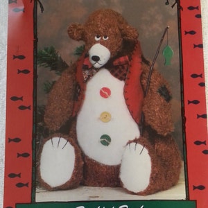 Puede incluir: Un patrón de artesanía para un oso de peluche llamado Bashful Buck. El patrón es para un oso marrón con vientre blanco y chaleco rojo. El oso sostiene una caña de pescar con un pez verde en el extremo. El patrón es de L.A. Designs y es el número 3002.