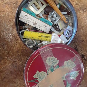May include: A red tin with a floral design and a white tape across the lid. The tin is open and filled with various small items, including buttons, pins, and a yellow ruler. The text on the label reads "Steel 100 Count Size S.G. Made in U.S.A. Pins".