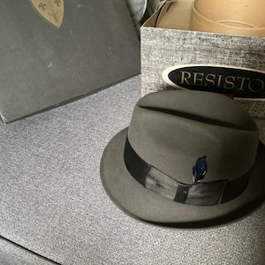 Op de afbeelding: Een grijze fedora hoed met een zwarte band en een kleine blauwe veer. De hoed staat op een grijze ondergrond naast een doos met de merknaam "RESISTOL". Een donkergrijze doos staat op de achtergrond.