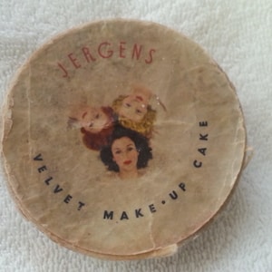 May include: Vintage Jergens Velvet Make-Up Cake compact with a white background and three women's faces in a triangle shape. The text "Jergens Velvet Make-Up Cake" is printed in black on the compact.