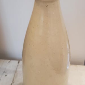 May include: A beige stoneware bottle with a rounded body and a narrow neck. The bottle has a smooth surface with small dark speckles. The opening is a lighter shade of beige. The bottle is likely an antique or vintage piece.