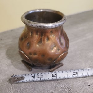 May include: A small, copper-coloured pot with a silver rim and three legs. The pot is approximately 5 cm tall and has a textured surface.