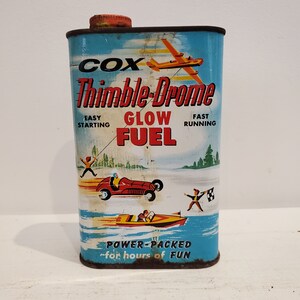 May include: Vintage tin can of Cox Thimble-Drome Glow Fuel for model aircraft. The can is blue with red, yellow, and white accents. It features a cartoon aeroplane, race car, and boat. The text on the can reads "Cox Thimble-Drome Glow Fuel, Easy Starting, Fast Running, Power-Packed for hours of fun."