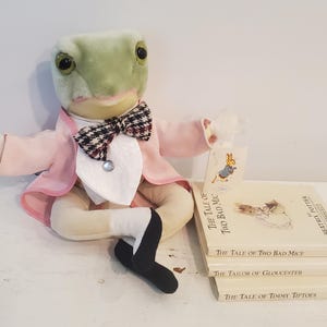 Peut inclure: Une grenouille en peluche portant une veste rose et un nœud papillon à carreaux noir et blanc est assise sur une surface blanche avec une pile de quatre livres. Les livres sont intitulés "The Tale of Two Bad Mice", "The Tale of Two Bad Mice", "The Tailor of Gloucester" et "The Tale of Timmy Tiptoes".