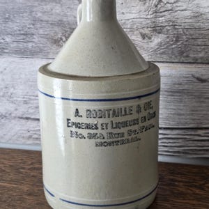 May include: A vintage stoneware jug with a handle and a conical top. The jug is a light beige colour with blue stripes and text that reads "A. ROBITAILLE & CIE EPICERIES ET LIQUEURS EN GROS No. 354 RUE ST. PAUL MONTREAL."