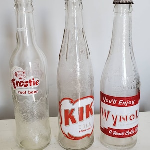 May include: Three vintage glass soda bottles. The bottles have labels with the text "Frostie root beer", "Kik Cola Serve Cold", and "Wynol A Real Cola Drink". The bottles are clear with red and white labels.