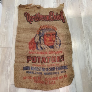 Peut inclure: Un sac en toile de jute brun avec le texte "Northern Chief" imprimé en noir. Le sac est décoré d'une illustration colorée d'un chef amérindien portant une coiffe. Le texte "Northern Grown Potatoes" est imprimé en bleu et rouge. Le sac comprend également le texte "Grown and Packed by John Bogestad & Son Farming, Donaldson, Minnesota 567, 100 lbs. Net Wt. 45.36 Kilos Net Wt. Produce of U.S.A."